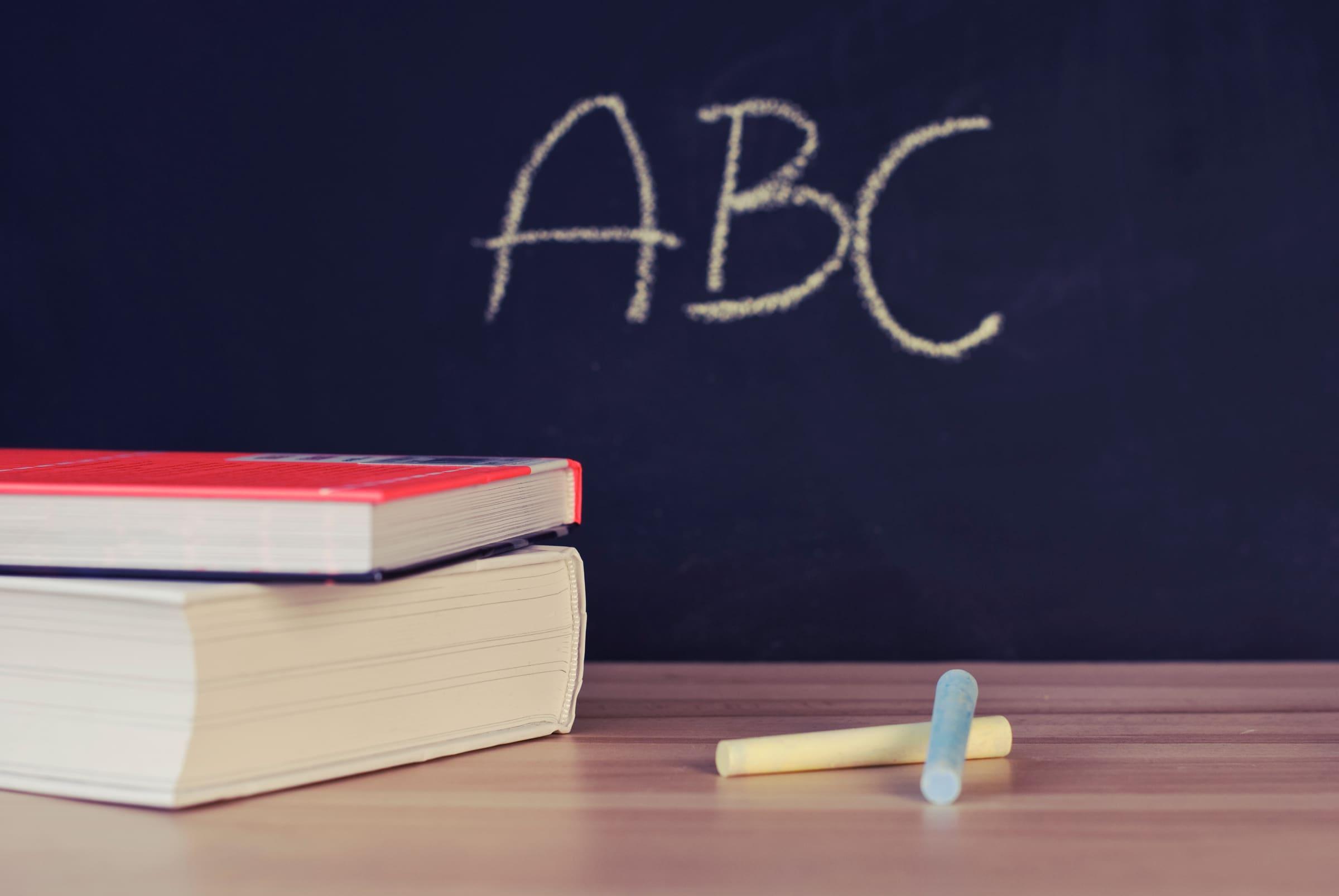 Des livres et des craies posés sur un bureau, devant un tableau noir où est écrit « ABC »